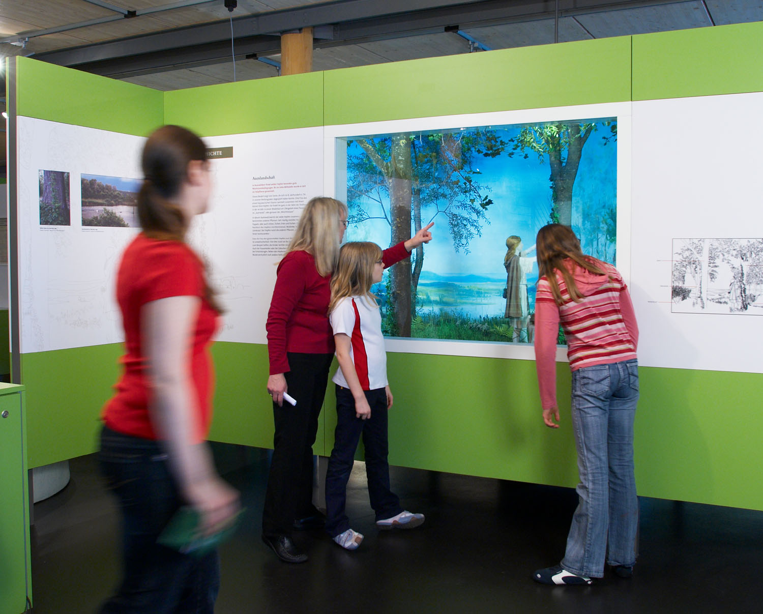 Die Besucherinnen stehen vor einer Vitrine. In der Vitrine sind Puppen. Die Puppen sind eine Frau und ihr Kind. Sie sammeln Hopfen. In einer Fluss-Landschaft. 